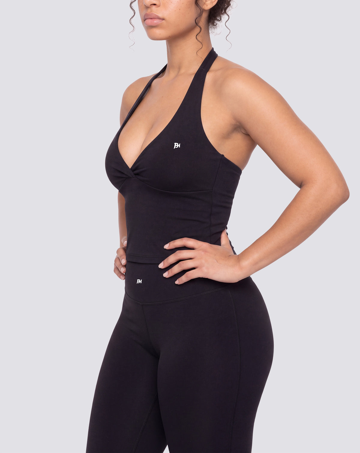 BEASTHAMMER Signature halter top in black, worn front three-quarter view with ruched twist and BH monogram, styled with matching Signature scrunch leggings