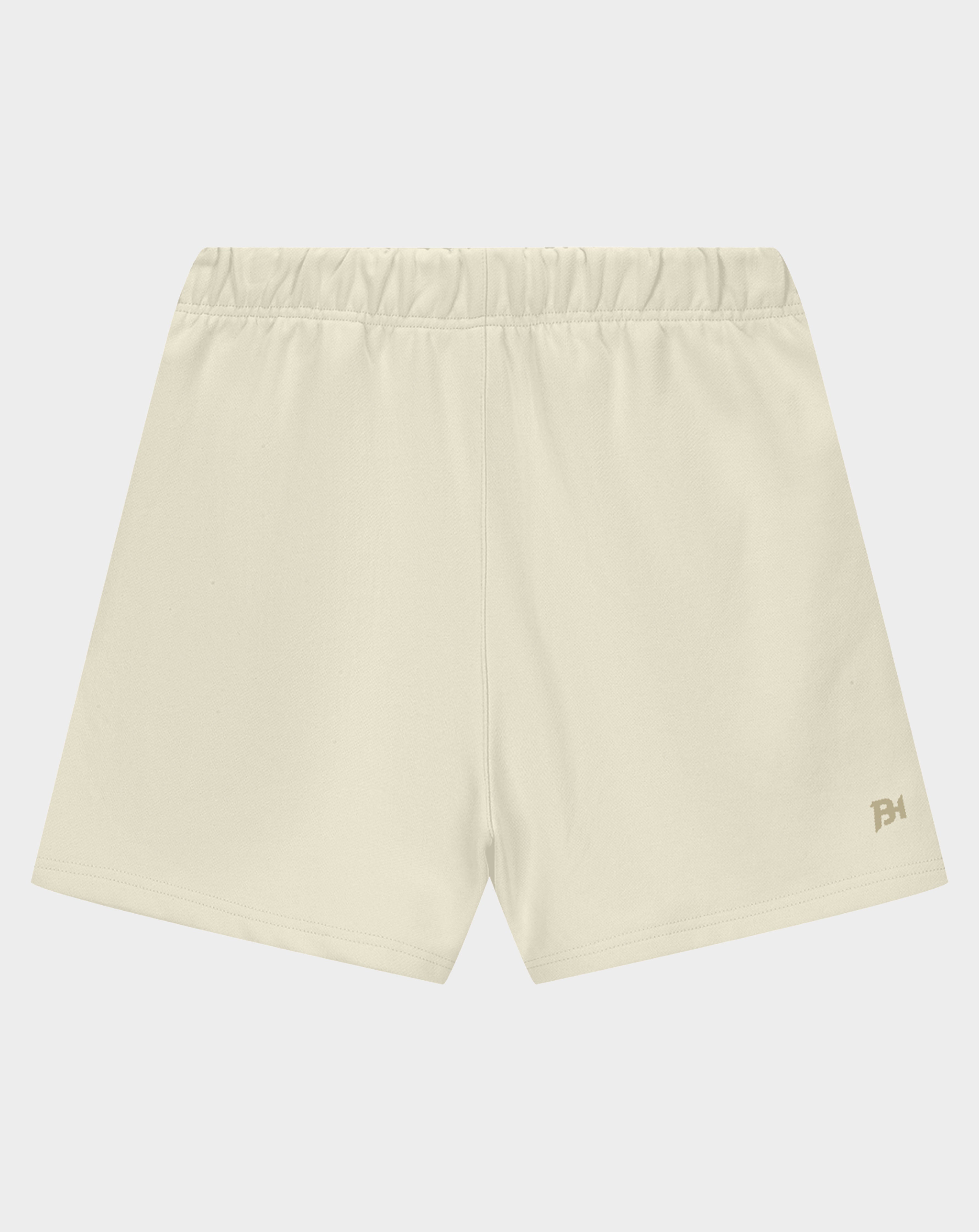 A pair of light cream athletic shorts with an elastic waistband and a small gold logo on the lower left hem, displayed against a light gray background.