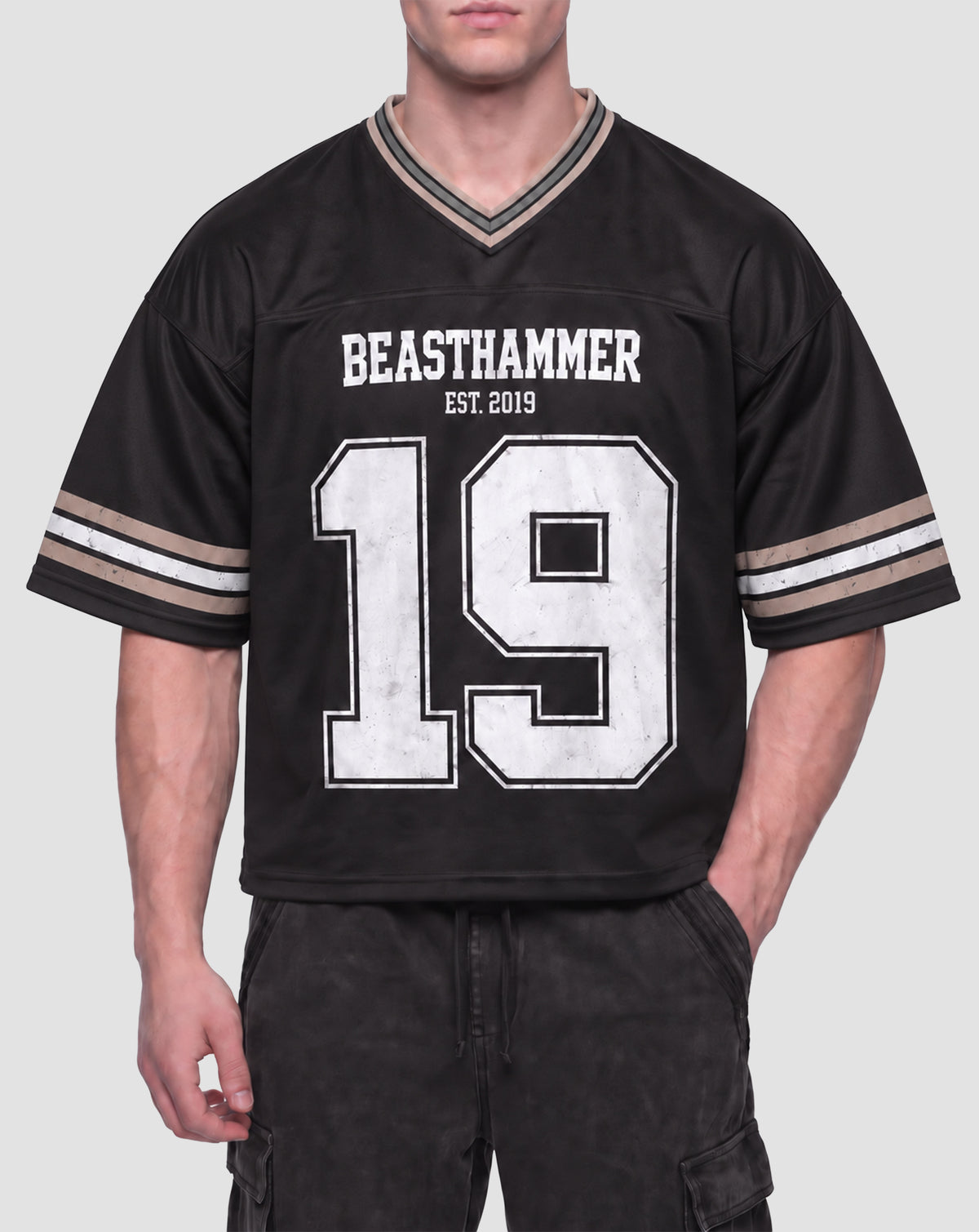 Man wearing the BEASTHAMMER Varsity 19 Jersey in black with black cargo trousers, front view with BEASTHAMMER and EST. 2019 above a distressed white 19, striped sleeves and V-neck