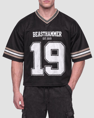 Man wearing the BEASTHAMMER Varsity 19 Jersey in black with black cargo trousers, front view with BEASTHAMMER and EST. 2019 above a distressed white 19, striped sleeves and V-neck