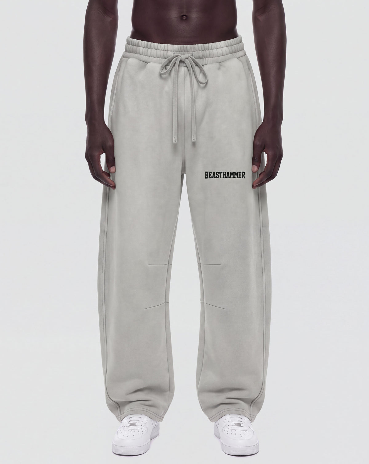 A model from the waist down wearing light gray sweatpants with a drawstring waist and the word 'BEASTAMMER' printed in black on the left thigh, paired with white sneakers against a light gray background.