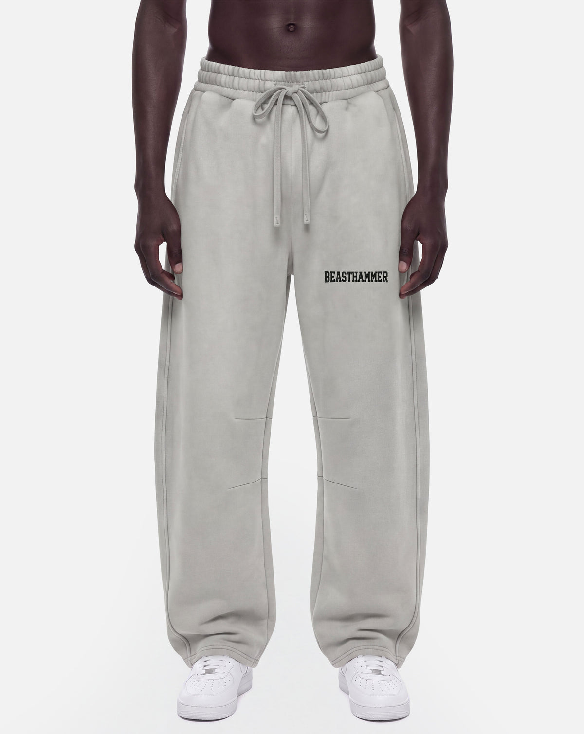 A model from the waist down wearing light gray sweatpants with a drawstring waist and the word 'BEASTAMMER' printed in black on the left thigh, paired with white sneakers against a light gray background.