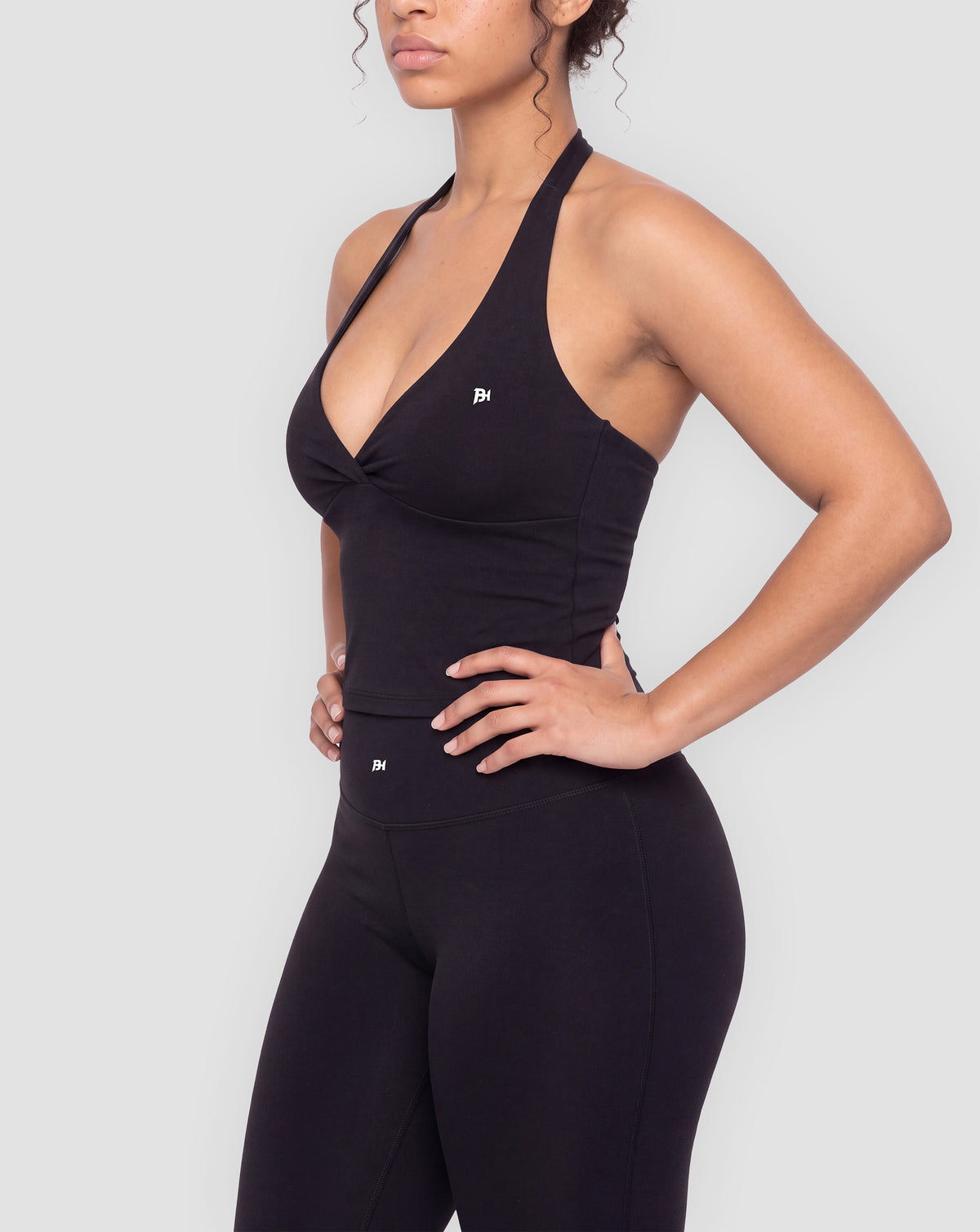 BEASTHAMMER Signature halter top in black, worn front three-quarter view with ruched twist and BH monogram, styled with matching Signature scrunch leggings