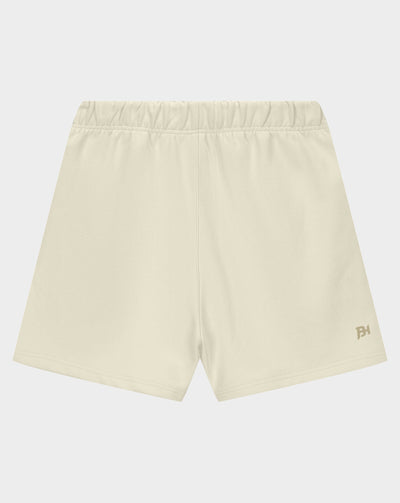 A pair of light cream athletic shorts with an elastic waistband and a small gold logo on the lower left hem, displayed against a light gray background.