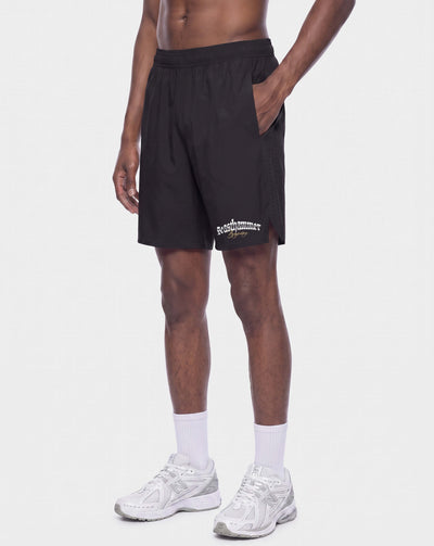 A model wearing black athletic shorts with a white embroidered logo on the lower left leg, paired with white crew socks and white sneakers, against a plain light gray background.
